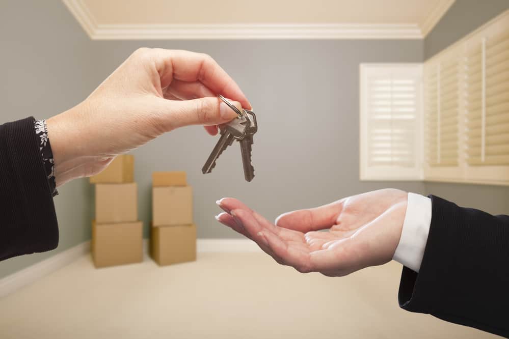 Woman Handing Over the House Keys To A New Home Inside Empty Grey Colored Room.