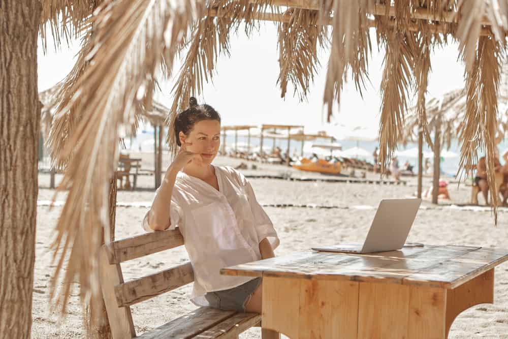 Pretty young woman using laptop in cafe on tropical beach in outdoor cafe. Work and travel